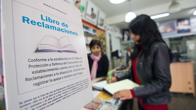 Presentamos la foto de un establecimiento comercial abierto al público. El local es pequeño, en el que una consumidora vestida de polo rojo manga larga y casaca negra, llena sobre un mostrador las hojas autocopiativas del libro de reclamaciones. Al frente de ella, pero un poco más hacia el fondo se encuentra la vendedora, apoyada con su codo en el mostrador. Está vestida de chompa de color negro y una bufanda multicolor sobre en el cuello.